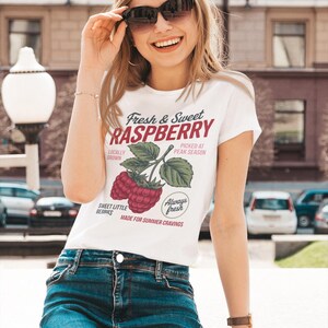 Op de afbeelding: Een wit t-shirt met korte mouwen voorzien van een afbeelding van frambozen aan een takje met de tekst Fresh & Sweet RASPBERRY, LOCALLY GROWN, PICKED AT PEAK SEASON, SWEET LITTLE BERRIES, Always fresh en MADE FOR SUMMER CRAVINGS in rood en zwart.