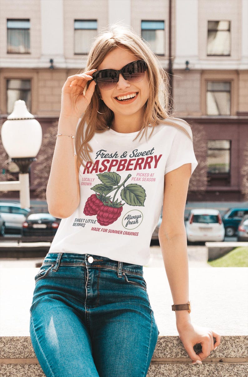 Op de afbeelding: Een wit t-shirt met korte mouwen voorzien van een afbeelding van frambozen aan een takje met de tekst Fresh & Sweet RASPBERRY, LOCALLY GROWN, PICKED AT PEAK SEASON, SWEET LITTLE BERRIES, Always fresh en MADE FOR SUMMER CRAVINGS in rood en zwart.
