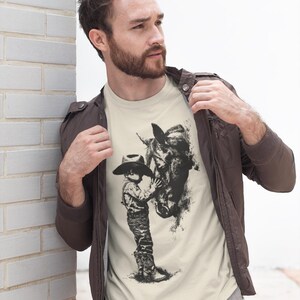 Puede incluir: Una camiseta de manga corta color crema con un estampado gr&aacute;fico negro estilo tinta que muestra a un ni&ntilde;o con sombrero de vaquero y botas de pie junto a un caballo. La camiseta se lleva debajo de una chaqueta marr&oacute;n oscuro y pantalones grises.