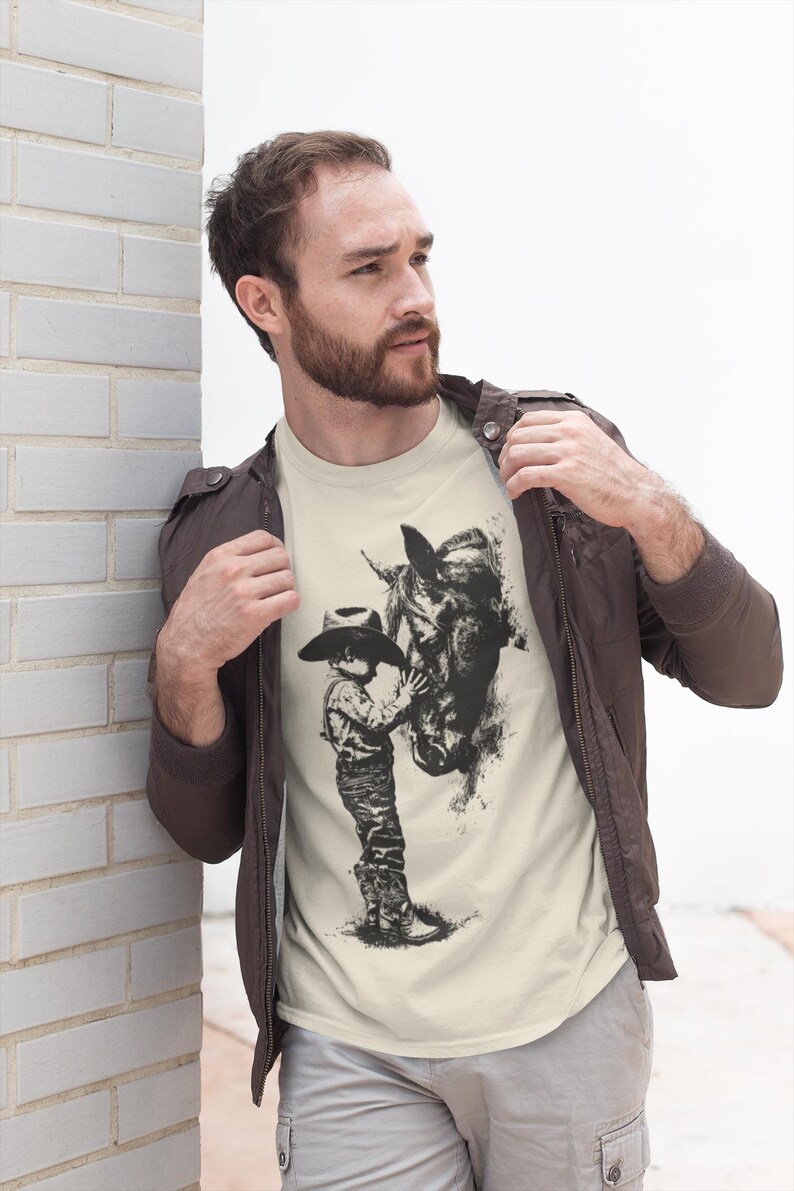 Puede incluir: Una camiseta de manga corta color crema con un estampado gr&aacute;fico negro estilo tinta que muestra a un ni&ntilde;o con sombrero de vaquero y botas de pie junto a un caballo. La camiseta se lleva debajo de una chaqueta marr&oacute;n oscuro y pantalones grises.