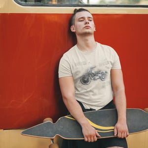 K&ouml;nnte beinhalten: Beige T-Shirt mit Motorrad-Design. Die Person tr&auml;gt schwarze Shorts und h&auml;lt ein Longboard. Der Hintergrund ist eine rote Wand.