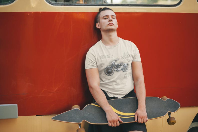 K&ouml;nnte beinhalten: Beige T-Shirt mit Motorrad-Design. Die Person tr&auml;gt schwarze Shorts und h&auml;lt ein Longboard. Der Hintergrund ist eine rote Wand.