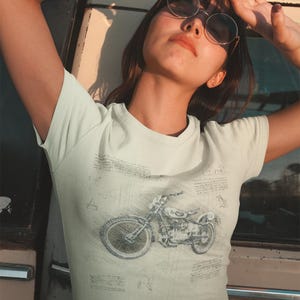 K&ouml;nnte beinhalten: Hellfarbenes T-Shirt mit einer detaillierten Skizze eines Motorrads. Das Design ist im Vintage-Stil gehalten, mit komplizierten Linien und Schattierungen. Das Shirt hat eine helle Farbe, m&ouml;glicherweise cremefarben oder cremefarben.