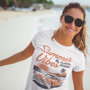 K&ouml;nnte beinhalten: Ein wei&szlig;es T-Shirt mit kurzen &Auml;rmeln und einem Grafikdruck eines Oldtimers an einem Strand bei Sonnenuntergang. Der Text Summer & Classic Rides ist in brauner Schrift &uuml;ber der Illustration des Autos, der Palme und der Sonne aufgedruckt.