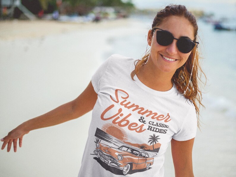 K&ouml;nnte beinhalten: Ein wei&szlig;es T-Shirt mit kurzen &Auml;rmeln und einem Grafikdruck eines Oldtimers an einem Strand bei Sonnenuntergang. Der Text Summer & Classic Rides ist in brauner Schrift &uuml;ber der Illustration des Autos, der Palme und der Sonne aufgedruckt.