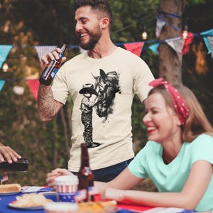 Puede incluir: Una camiseta de manga corta color crema con un estampado gr&aacute;fico negro estilo tinta que muestra a una persona con sombrero de vaquero junto a un caballo. La camiseta tiene cuello redondo y un corte est&aacute;ndar con una ilustraci&oacute;n detallada.
