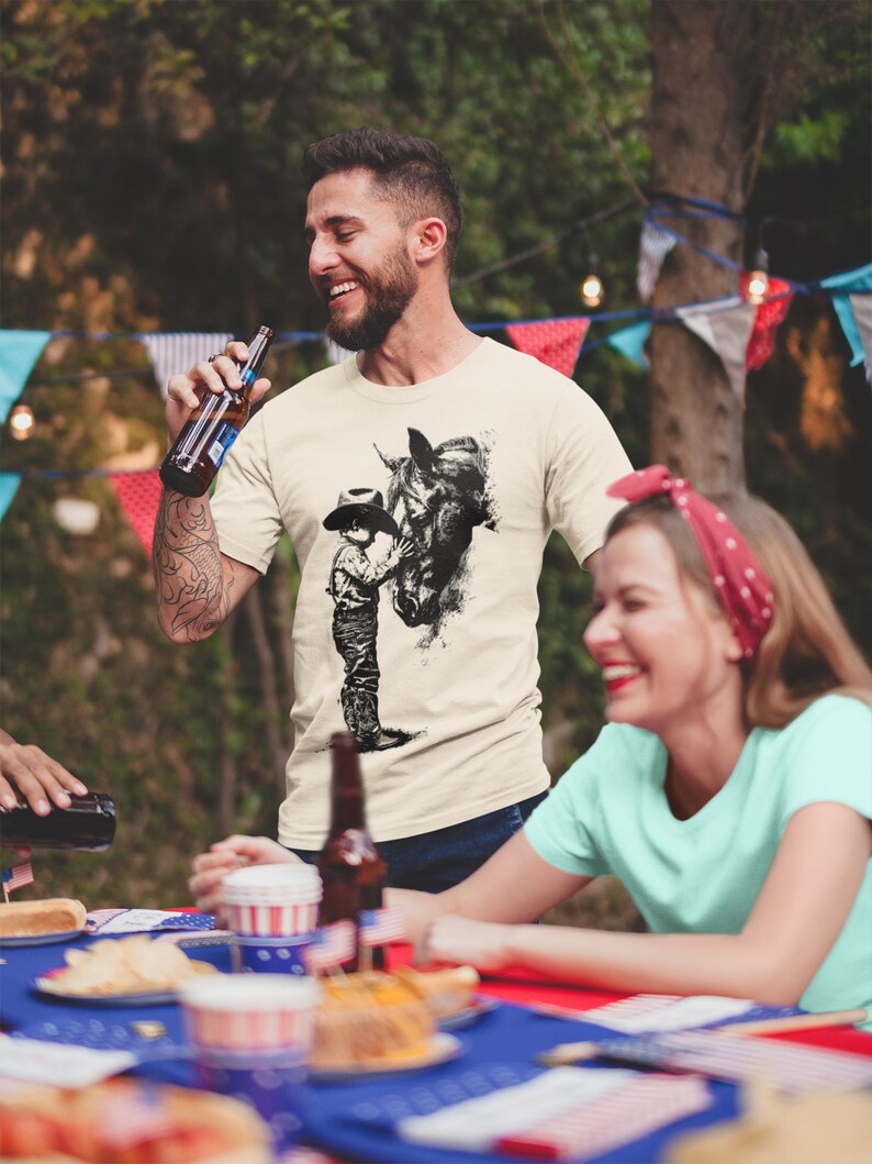 Puede incluir: Una camiseta de manga corta color crema con un estampado gr&aacute;fico negro estilo tinta que muestra a una persona con sombrero de vaquero junto a un caballo. La camiseta tiene cuello redondo y un corte est&aacute;ndar con una ilustraci&oacute;n detallada.