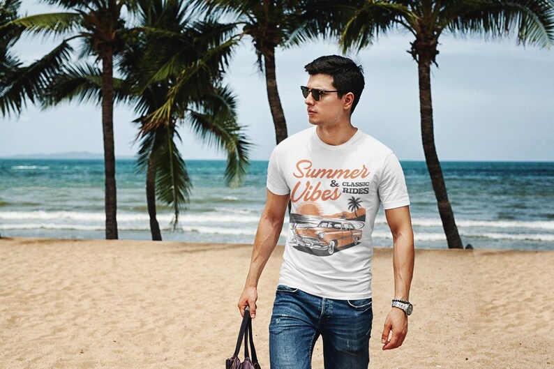 K&ouml;nnte beinhalten: Ein wei&szlig;es kurz&auml;rmeliges T-Shirt mit einem Grafikdruck eines Oldtimers am Strand mit Palmen und dem Text Summer Vibes & Classic Rides in orangefarbener und schwarzer Schrift, kombiniert mit einer blauen Jeans und einer dunklen Sonnenbrille.