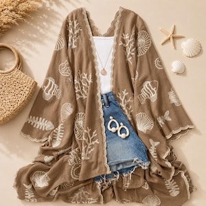 May include: A brown open-front kimono featuring a white embroidered pattern of seashells, coral, and fish. The garment has scalloped edges and is layered over a white tank top, blue denim shorts, and a gold necklace with a round pendant.