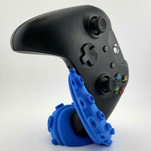 May include: A black wireless video game controller resting on a bright blue 3D-printed stand shaped like an octopus tentacle. The controller features a directional pad, two analogue sticks, and four coloured action buttons labelled A, B, X, and Y.