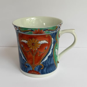 May include: A ceramic mug featuring a vibrant, multi-colored pattern with a prominent orange floral motif centered on a deep blue background. The design includes green accents, white scrollwork, and a white interior with a matching white handle.