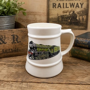 May include: A white ceramic mug featuring a detailed illustration of a green steam locomotive train on the side. The mug has a wide base, a cylindrical body with two horizontal raised ridges, and a curved handle on the right side.
