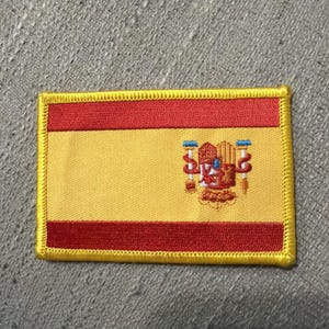Può includere: Una toppa rettangolare ricamata raffigurante la bandiera della Spagna. Il design è composto da tre strisce orizzontali rosse e gialle, con lo stemma spagnolo centrato sulla striscia gialla. L'intera toppa presenta un bordo cucito giallo.