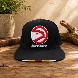 Puede incluir: Una gorra de béisbol negra con el logotipo de los Atlanta Hawks bordado en rojo y blanco en la parte delantera. El texto Atlanta Hawks está cosido en blanco debajo del logo. La visera plana tiene una fina franja horizontal roja, amarilla y blanca.