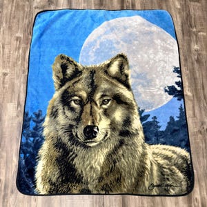 Op de afbeelding: Een rechthoekige pluche deken met een gedetailleerde afbeelding van een wolf op de voorgrond. De achtergrond toont een grote, heldere volle maan tegen een diepblauwe nachtelijke hemel met donkere silhouetten van dennenbomen onderaan.