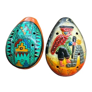May include: Two egg-shaped ceramic ocarinas. The left ocarina features a bright orange base with a teal, yellow, and black geometric pattern. The right ocarina displays a painted figure in a red hat and cloak, standing near a stone structure.