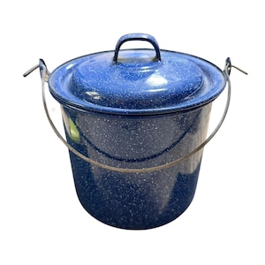 Op de afbeelding: Een cilindrische blauwe geëmailleerde emmer met een wit gespikkeld patroon, voorzien van een bijpassend deksel met een centrale handgreep en een dunne, gebogen metalen hengsel aan de zijkanten om te dragen. De afwerking is glanzend en glad.