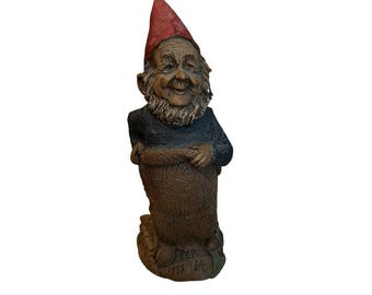 Könnte beinhalten: Eine Zwergenfigur mit einer spitzen roten Mütze, einem dunkelblauen langärmeligen Oberteil und einer braunen Hose. Die Figur hat einen strukturierten Bart und ein lächelndes Gesicht. Der Sockel zeigt den geprägten Text "CEP 1787 64".