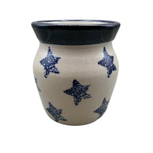 Może przedstawiać: Ceramiczny garnek o nakrapianym, kremowym korpusie z wzorem w ciemnoniebieskie gwiazdy. Naczynie posiada szeroki, rozszerzający się otwór z jednolitym ciemnoniebieskim brzegiem. Powierzchnia ma gładkie, szkliwione wykończenie i zaokrąglony kształt.