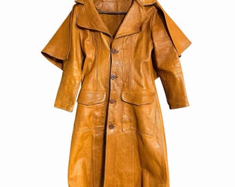 Puede incluir: Una gabardina de cuero color canela con cuello ancho, cierre frontal de botones y una capa superpuesta sobre los hombros. El abrigo incluye dos bolsillos delanteros con solapa y mangas largas, mostrando un acabado de cuero natural texturizado.