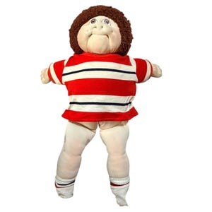 May include: A soft fabric doll with a round face, brown yarn hair, and brown eyes. The doll wears a short-sleeved shirt featuring horizontal red, white, and dark blue stripes, paired with plain off-white fabric legs and white socks with red and blue trim.