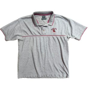 Könnte beinhalten: Ein kurzärmeliges Poloshirt in Grau meliert mit weinroten Akzenten am Kragen, an den Ärmelbündchen und einem horizontalen Bruststreifen. Das Arizona State Logo ist in Weinrot und Gold auf der linken Brust gestickt. Mit drei Knöpfen.