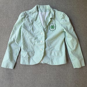 Pode incluir: Um blazer às riscas verde-claro e branco com lapela entalhada, três botões frontais e dois bolsos com aba. Um emblema branco em forma de escudo com um trevo verde de quatro folhas e a letra H em cada folha está cosido no peito.