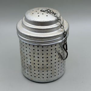 May include: A cylindrical metal tea infuser with a perforated body and a matching lid. The lid features a small handle loop attached to a metal chain that ends in a hook. The surface of the silver-toned metal shows signs of wear and light scratches.