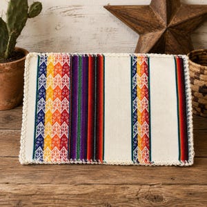May include: A rectangular fabric placemat featuring a cream-colored base with vertical stripes in vibrant shades of orange, red, purple, green, and blue. The design includes intricate geometric patterns and a decorative cream crochet border.