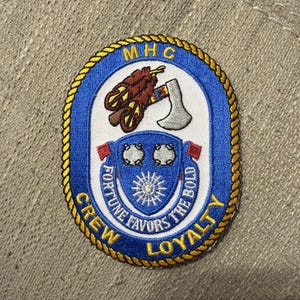May include: An oval embroidered patch with a blue border and yellow rope trim. It features a brown feather, a silver axe, and a blue shield with two silver circles and a sunburst. Text includes MHC, CREW, LOYALTY, and FORTUNE FAVORS THE BOLD.