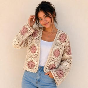 May include: A long-sleeved crochet cardigan constructed from square motifs in shades of cream and dusty rose. The garment features an open front design with a scalloped edge finish, showcasing a repeating floral pattern throughout the knit fabric.