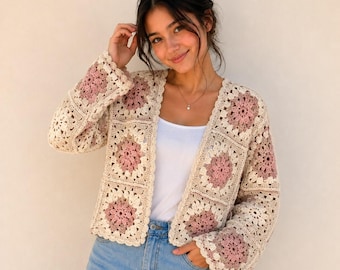 May include: A long-sleeved crochet cardigan constructed from square motifs in shades of cream and dusty rose. The garment features an open front design with a scalloped edge finish, showcasing a repeating floral pattern throughout the knit fabric.