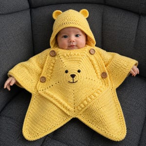 May include: A bright yellow crocheted baby poncho featuring a hood with small bear ears. The garment has a star-shaped design with a central bear face embroidery, textured bobble stitch details, and four brown wooden buttons on the front.