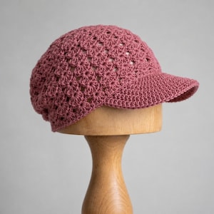 Peut inclure: Une casquette en crochet rose poudré présentant un motif répétitif en forme d'étoile sur la calotte et une visière solide aux mailles serrées. Le chapeau est réalisé avec un travail de fil complexe, affichant un design texturé et ajouré.
