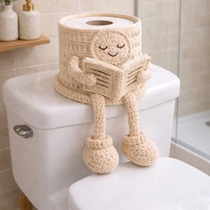 May include: A beige crochet toilet paper roll cover featuring a character with a smiling face holding an open book. The cover has a textured grid pattern, dangling legs with rounded feet, and arms attached to the book, designed to fit over a standard roll.