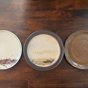 May include: Three circular ceramic plates arranged in a row. The left plate is off-white with brown drip patterns at the bottom. The center plate features a tan center with a dark brown rim and textured brown accents. The right plate is solid brown.