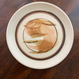 May include: A round ceramic plate with a speckled off-white base. The center features a circular design with abstract, sweeping brushstrokes in shades of tan and brown, enclosed by a dark brown ring that frames the central artistic motif.
