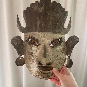 May include: A weathered metal wall mask featuring a tall, crown-like headdress, prominent ears, and dangling circular earrings. The face has a textured, aged gray finish with dark brown accents, defined eyes with dark lashes, and a distinct nose and lips.