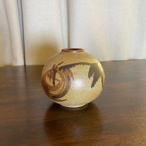 May include: A spherical ceramic vase with a tan, speckled glaze and dark brown, abstract brushstroke patterns. The vase features a small, circular opening at the top with a smooth, dark brown rim, showcasing a rounded body and a flat base.