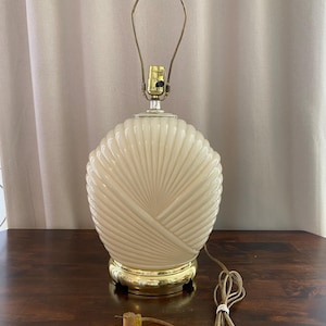 May include: A table lamp featuring a cream-colored, oval-shaped glass body with a textured, radiating ribbed pattern. The lamp sits on a polished gold-tone metal base and includes a gold-tone metal harp fixture at the top with a brown electrical cord.
