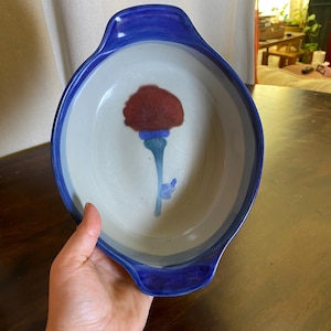 May include: An oval ceramic baking dish with a glossy cobalt blue rim and handles. The interior is a light grey, featuring a hand-painted floral motif with a dark red flower head and a blue stem. The dish has a smooth, glazed finish throughout.