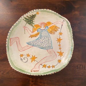 May include: A round ceramic plate with an irregular, textured edge featuring a hand-painted illustration of a winged figure in a blue polka-dot dress. The figure holds a green Christmas tree and a string of yellow stars. The name Chris is signed.