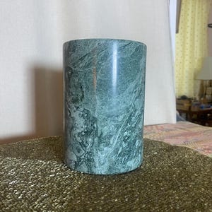 May include: A cylindrical vessel crafted from polished green marble, featuring natural white veining throughout the stone surface. The object has a smooth, vertical exterior and a flat base, showcasing the deep forest green color and intricate patterns.