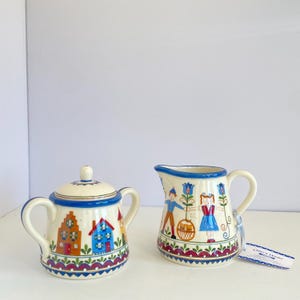 May include: A ceramic sugar bowl with a lid and a matching creamer pitcher, both featuring hand-painted designs of colorful houses, flowers, and figures. The pieces have blue rims and decorative patterns along the base, with a small tag labeled Oma's House.