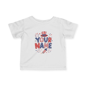 Puede incluir: Una camiseta blanca de manga corta con un estampado gráfico que muestra el texto YOUR NAME en patrones rojos, blancos y azules. El diseño incluye un sombrero de copa, un cohete de fuegos artificiales y estrellas en la parte frontal.