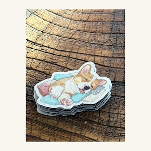 Può includere: Una pila di adesivi fustellati con un'illustrazione in stile ricamo di un cucciolo di Corgi che dorme. Il cucciolo ha il pelo marrone chiaro e bianco, il naso nero e riposa su un cuscino azzurro con un piccolo cuscino rosa vicino.
