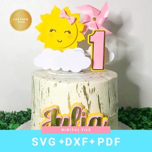 May include: A cake topper featuring a yellow sun with a pink bow, white clouds, a pink pinwheel, and a pink number one. The name Julia is displayed in gold and pink lettering. The topper is shown on a white cake with gold paint splatters.