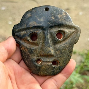 May include: A dark green, mottled stone pendant carved into a stylised face shape. The piece features two large, almond-shaped eye cutouts, two smaller mouth cutouts, and a single hole at the top for suspension. The surface has a smooth, polished texture.