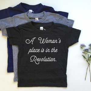 May include: Three t-shirts stacked on top of each other. The top shirt is black with white text that reads "A Woman's place is in the Revolution."