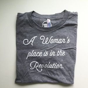 May include: A heather grey t-shirt with white text that reads "A Woman's place is in the Revolution."